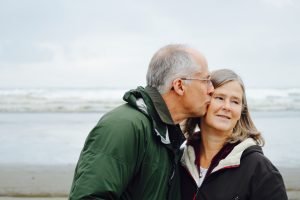 older man kissing older woman on check beside body of water