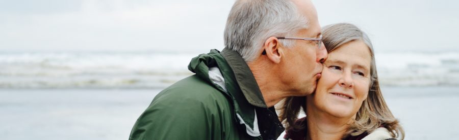older man kissing older woman on check beside body of water